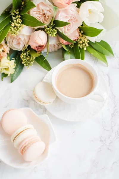 Elegante zoete dessert Macarons, kopje koffie en pastelkleurige beige bloemen boeket op wit marmer — Stockfoto