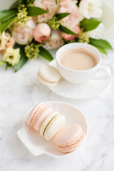 Elegant sweet dessert macarons, cup of coffee and pastel colored beige flowers bouquet on white marble — Stock Photo, Image