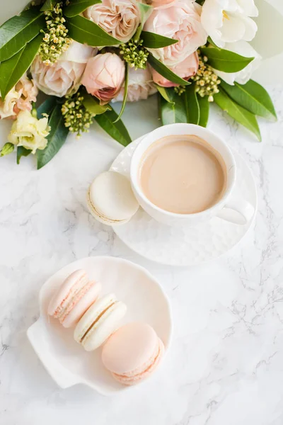 Macarons de sobremesa doce elegante, xícara de café e pastel colorido buquê de flores bege em mármore branco — Fotografia de Stock