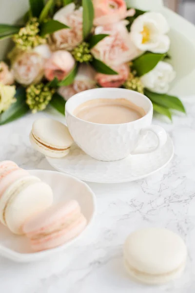 Elegant sweet dessert macarons, cup of coffee and pastel colored beige flowers bouquet on white marble — Stock Photo, Image