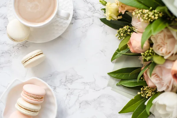 Elegant sweet dessert macarons, cup of coffee and pastel colored beige flowers bouquet on white marble — Stock Photo, Image