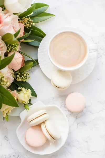 Κομψό γλυκό επιδόρπιο μακαρόνια, φλιτζάνι καφέ και παστέλ χρώματα μπεζ λουλούδια μπουκέτο σε λευκό μάρμαρο — Φωτογραφία Αρχείου