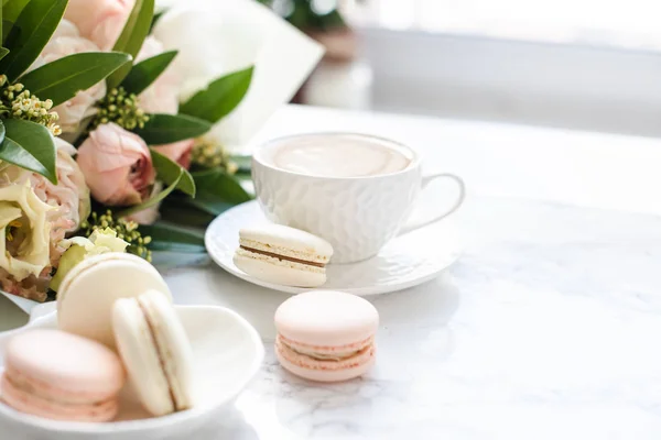 Elegante zoete dessert Macarons, kopje koffie en pastelkleurige beige bloemen boeket op wit marmer — Stockfoto