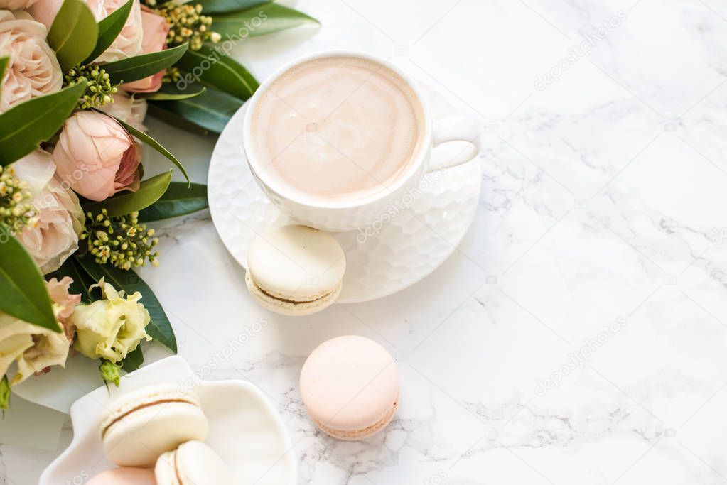 Elegant sweet dessert macarons, cup of coffee and pastel colored beige flowers bouquet on white marble