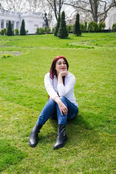 Ung kvinna i vit skjorta och Blå jeans sitter på ett grönt gräs — Stockfoto