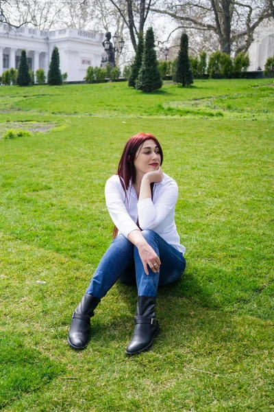 白いシャツと青いジーンズの若い女性が緑の芝生の上に座っている — ストック写真