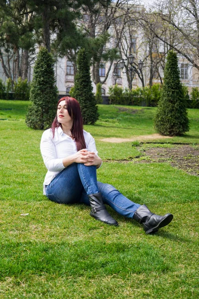 Mladá žena v bílé košili a modré džínách, které sedí na zelené trávě — Stock fotografie