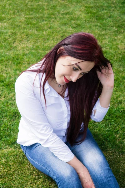Jonge vrouw in wit overhemd en blauwe jeans zittend op een groen gras — Stockfoto