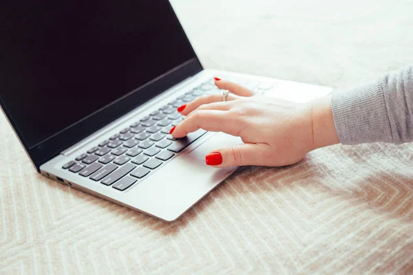 Womans hand with red nails on laptop keyboard, lady using laptop on bed blanket