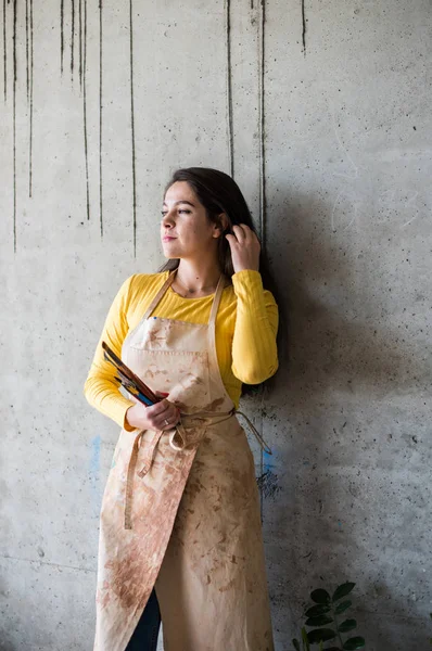 Young beautiful lady artist in apron with paint stains in her loft artistic studio