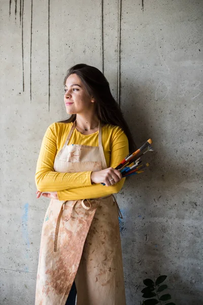Junge schöne Künstlerin in Schürze mit Farbflecken in ihrem künstlerischen Atelier auf dem Dachboden — Stockfoto