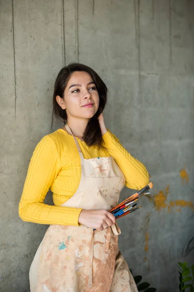 Young beautiful lady artist in apron with paint stains in her loft artistic studio