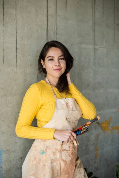 Junge schöne Künstlerin in Schürze mit Farbflecken in ihrem künstlerischen Atelier auf dem Dachboden — Stockfoto