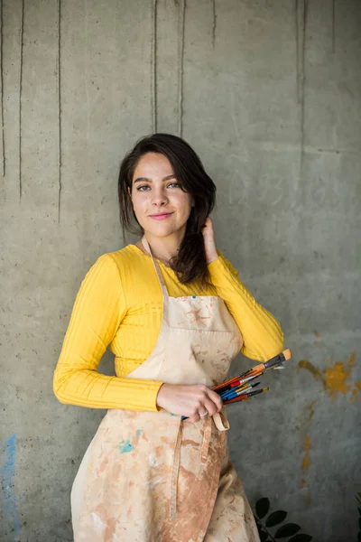 Young beautiful lady artist in apron with paint stains in her loft artistic studio