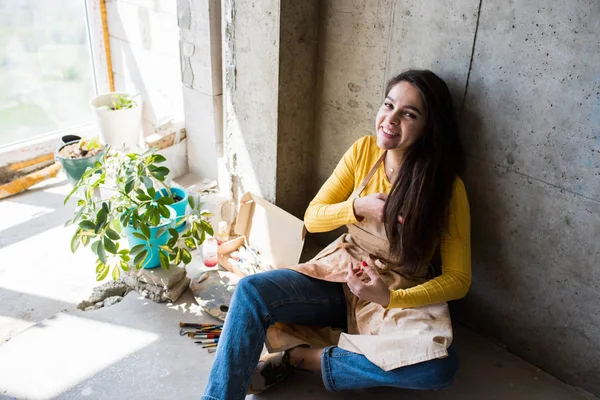 Joven hermosa artista en delantal con manchas de pintura sentado en el suelo en su estudio —  Fotos de Stock