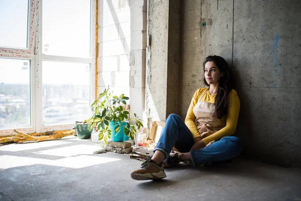 Jonge mooie dame artiest in schort met verf vlekken zitten op de vloer in haar studio — Stockfoto