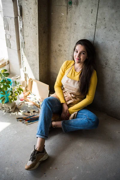 Jovem artista bonita senhora em avental com manchas de tinta sentado no chão em seu estúdio — Fotografia de Stock