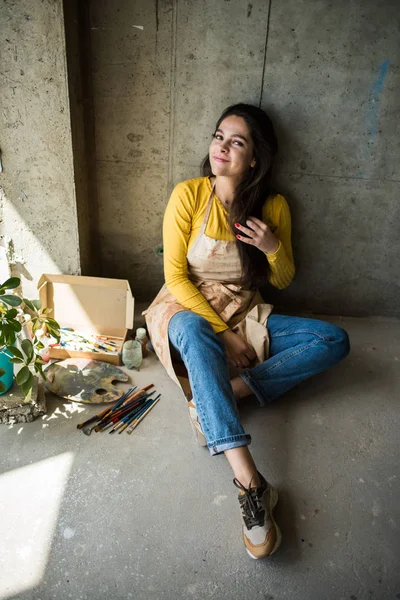 Jonge mooie dame artiest in schort met verf vlekken zitten op de vloer in haar studio — Stockfoto