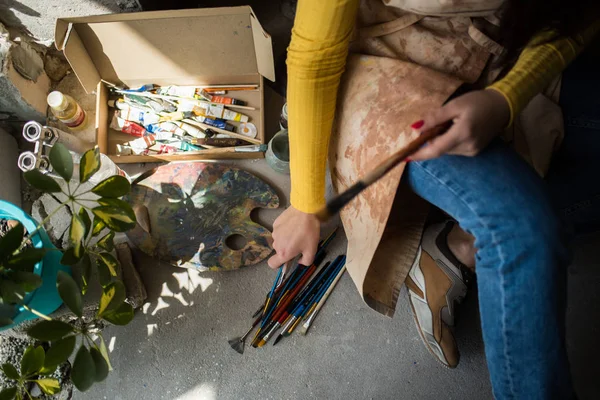 Giovane bella signora artista in grembiule con macchie di vernice seduta sul pavimento nel suo studio — Foto Stock