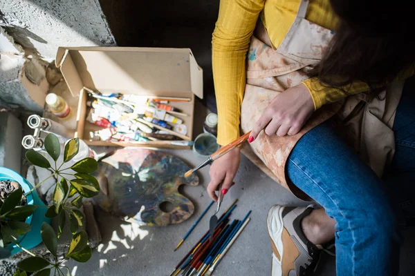 Молодая красивая художница в фартуке с пятнами от краски, сидящая на полу в своей студии — стоковое фото