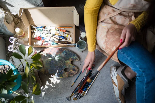 Joven hermosa artista en delantal con manchas de pintura sentado en el suelo en su estudio —  Fotos de Stock