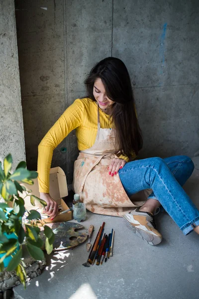 Jeune belle dame artiste dans le tablier avec des taches de peinture assis sur le sol dans son atelier — Photo