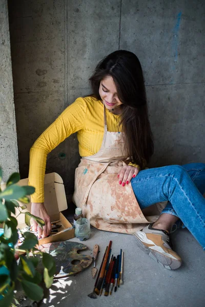 Jeune belle dame artiste dans le tablier avec des taches de peinture assis sur le sol dans son atelier — Photo