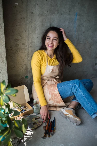 Jeune belle dame artiste dans le tablier avec des taches de peinture assis sur le sol dans son atelier — Photo