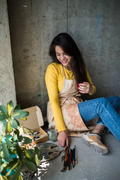 Jeune belle dame artiste dans le tablier avec des taches de peinture assis sur le sol dans son atelier — Photo