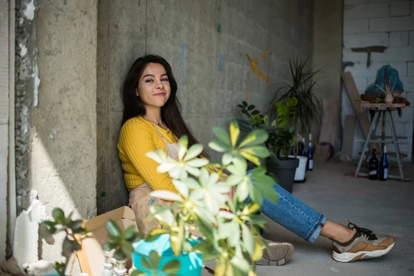 Joven hermosa artista en delantal con manchas de pintura sentado en el suelo en su estudio —  Fotos de Stock