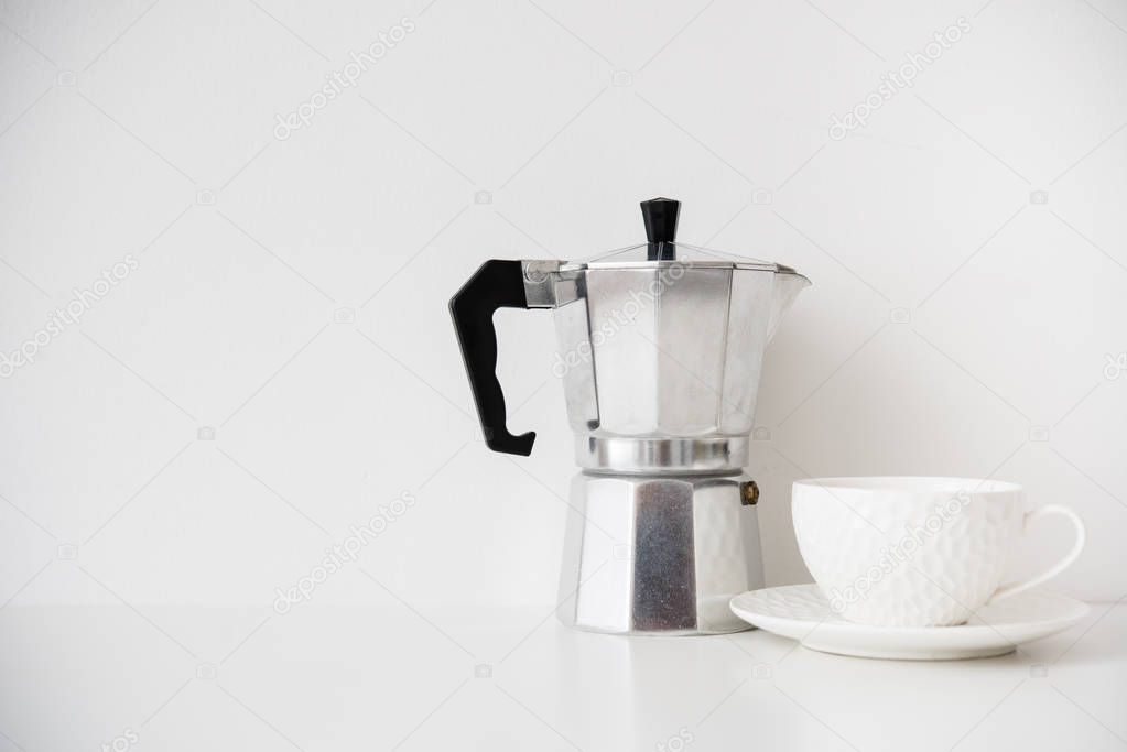 Metal coffee maker and white porcelain cup on table with blank wall