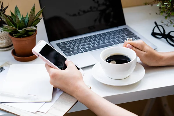 Frauenhand mit Tasse Kaffee und Smartphone im gemütlichen Büro mit Laptop und grünen Pflanzen — Stockfoto