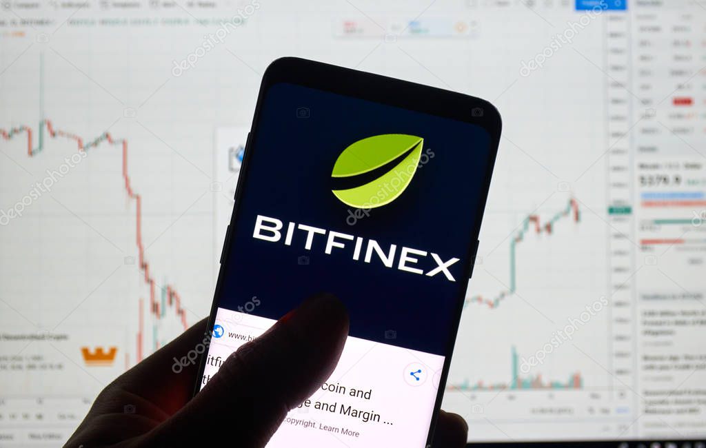 MONTREAL, CANADA - APRIL 26, 2019: Bitfinex cryptocurrency exchange logo and application on Android Samsung Galaxy s9 Plus screen in a hand over a laptop display with bitcoin chart on it.
