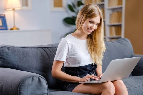 Mujer bonita usando el ordenador portátil sentado en el sofá acogedor —  Fotos de Stock