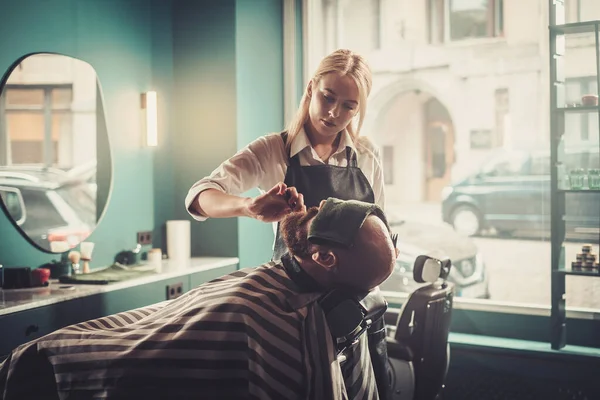 Hipster Man senta-se na cadeira com os olhos fechados e uma toalha durante o corte profissional da barba . — Fotografia de Stock