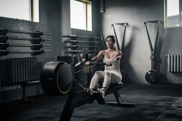 Mujer en ropa deportiva blanca tiene entrenamiento en la máquina de remo . — Foto de Stock