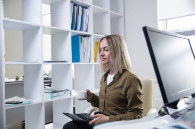 Genç bir kadın sabahları ofiste kahve içer ve hesap makinesiyle hesaplamalar yapar..