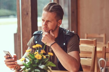 Bir şık saç ve sakal dalgın hipster yol kenarına yerleştirilen bir kafede bir masada oturuyor, bir akıllı telefon tutar.