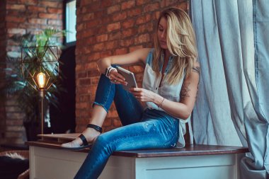 Bir çatı katı iç bir stüdyoda bir tuğla duvara bir masada otururken bir tablet moda sarışın kadın blogger tutar.