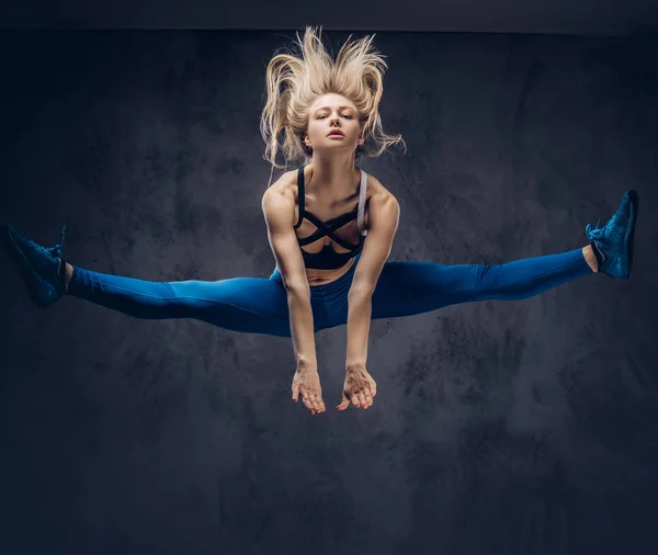 Jonge blonde ballerina in sportkleding dansen en springt in een studio. Geïsoleerd op een donkere achtergrond. — Stockfoto