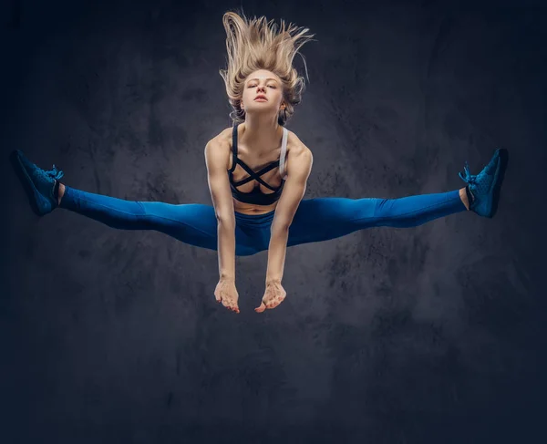 Jonge blonde ballerina in sportkleding dansen en springt in een studio. Geïsoleerd op een donkere achtergrond. — Stockfoto