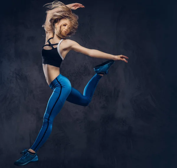 Young blonde ballerina in sportswear dances and jumps in a studio. Isolated on a dark background. — Stock Photo, Image