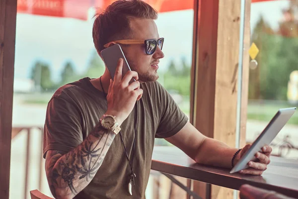 Όμορφος μοντέρνος hipster κάθεται σε ένα τραπέζι σε ένα καφέ άκρη του δρόμου, μιλώντας στο τηλέφωνο και κατέχει το δισκίο. — Φωτογραφία Αρχείου