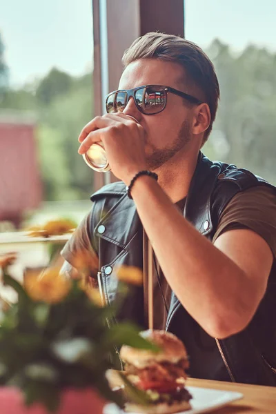 Hipster avec une coiffure élégante et la barbe est assis à une table, arrêté pour déjeuner dans un café en bord de route . — Photo