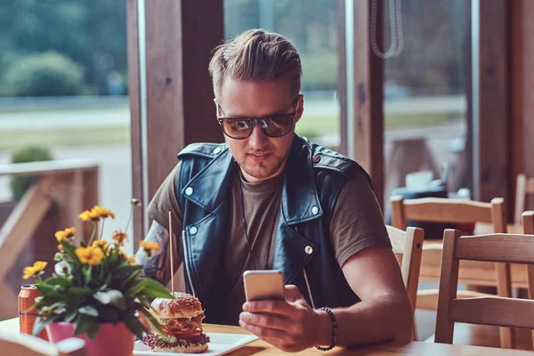 Hipster met een stijlvol kapsel en baard zit aan een tafel, met behulp van een smartphone tijdens de lunch in een cafe langs de weg. — Stockfoto