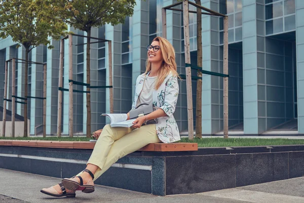 Encantadora rubia vestida de mujer moderna, estudiando con un libro, sentada en un banco en el parque contra un rascacielos . —  Fotos de Stock