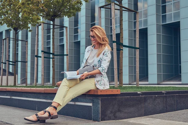 Encantadora rubia vestida de mujer moderna, estudiando con un libro, sentada en un banco en el parque contra un rascacielos . —  Fotos de Stock