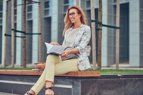 Charmiga blond kvinna i moderna kläder, studera med en bok, sitter på en bänk i parken mot en skyskrapa. — Stockfoto