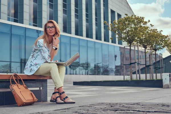 Stylische Modebloggerin, die ihren Laptop zur Arbeit benutzt, während sie draußen auf einer Bank vor einem Wolkenkratzer sitzt. — Stockfoto