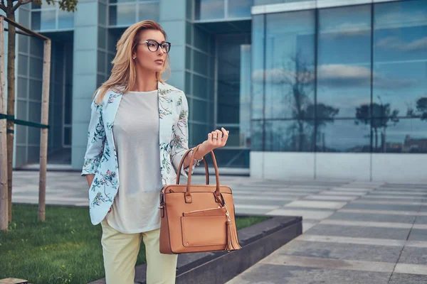 Красивая модная женщина в стильной одежде и очках с сумочкой, стоящая напротив небоскреба . — стоковое фото